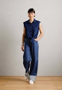 Chemise sans manches bleu marine à boutons, associée à un pantalon en denim bleu ample avec une jambe large et des ourlets retroussés, complétée par des baskets blanches.
