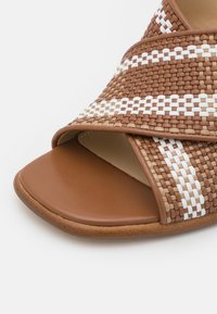 Sandale marron à bout ouvert avec de larges brides tissées en marron, blanc et beige qui se croisent sur le pied.