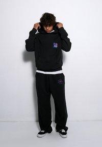 Czarna bluza z kapturem, posiadająca kieszeń z przodu i kolorowe logo. W połączeniu z dopasowanymi czarnymi spodniami dresowymi, które mają graficzne akcenty.
