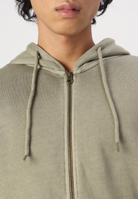 Hellgrüne Zip-Hoodie aus weichem, strukturiertem Material. Besitzt eine Kapuze, Kordeln und einen metallischen Reißverschluss, der glatte, neutrale Töne zeigt.
