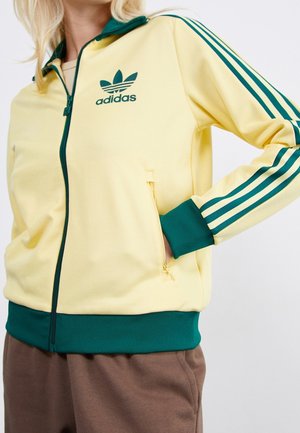 Person iført en gul Adidas lynlåsjakke med mørkegrønne striber, manchetter og linning, hånd i jakkelommen, brune bukser synlige.