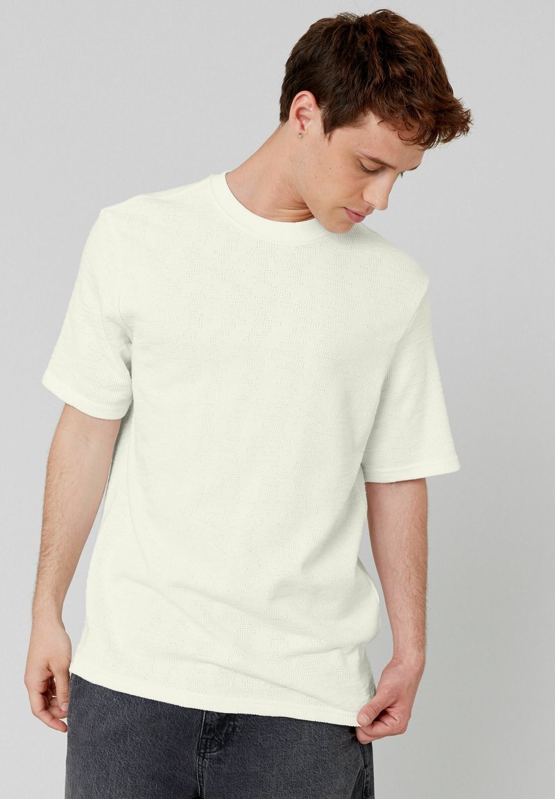 T-shirt blanche à manches courtes avec une surface texturée. Col rond, coupe décontractée, associée à un jean gris foncé. Design minimaliste, sans motifs visibles.