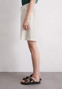 Pantalones cortos de lino en beige claro, con una cintura elástica y un cordón ajustable. Combinados con sandalias negras con acentos metálicos.