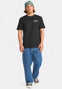T-shirt preto de manga curta com logótipo no lado esquerdo do peito, combinado com jeans azuis de corte largo e ténis pretos e brancos.