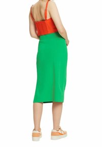 Femme portant un haut sans manches orange vif, une jupe verte au genou et des baskets orange-blanc, debout, dos face à l’avant.