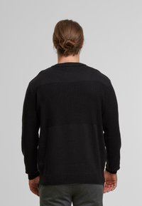Schwarzer Strickpullover mit strukturiertem Muster, der einen einfarbigen Bereich im oberen Teil des Körpers sowie gerippte Bündchen und Saum aufweist.