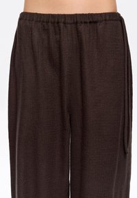 Pantalones marrones, texturizados, con cintura elástica y corte relajado, que presentan un detalle de lazo en el lateral y un sutil patrón de canalé vertical.