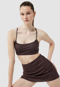 Młoda kobieta pozująca w brązowej bluzce crop top w kropki oraz dopasowanej spódnicy z cienkimi ramiączkami na neutralnym tle.