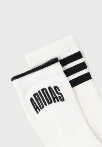 Άσπρες ραβδωτές κάλτσες με μαύρες λεπτομέρειες. Μία κάλτσα έχει το λογότυπο "ADIDAS" με μαύρα γράμματα· η άλλη έχει τρεις μαύρες ρίγες κοντά στην κορυφή.
