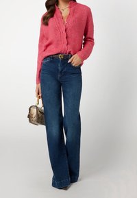 Roze gebreide cardigan met golvende randen, gecombineerd met blauwe flare jeans. Accessoire is een kleine handtas met een slangenhuidpatroon.