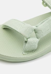 Sandalia verde claro con una suela texturizada, que presenta amplias correas de tela aseguradas con herrajes redondos y un diseño suave y flexible.
