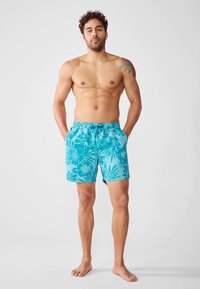 Άρρεν χωρίς πουκάμισο με ανοιχτόχρωμα μπλε μαγιό με λουλούδια στέκεται ξυπόλητος σε απλό ανοιχτό φόντο, με τα χέρια στις τσέπες, δείχνοντας ένα τατουάζ στον ώμο.