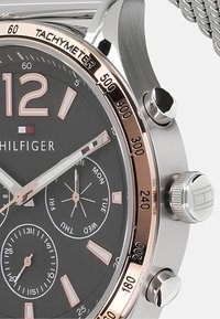 Montre chronographe argentée avec un cadran noir, des accents en or rose, une lunette tachymétrique et un bracelet en métal texturé. Présente des marqueurs de jour de la semaine et des sous-cadrans.