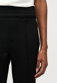 Κοντινό πλάνο ενός ατόμου που φοράει μαύρο παντελόνι με ψηλή μέση και λευκό τοπ, με το δεξί του χέρι χαλαρό στο πλάι.