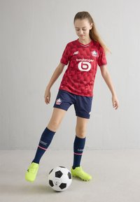 Röd fotbollströja med 3D-geometriskt mönster, marinblå shorts, blå knästrumpor, lysande gula fotbollsskor och en svartvit fotboll.