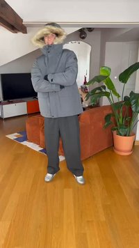 Parka grigia isolata con cappuccio foderato di pelliccia, abbinata a pantaloni larghi scuri e sneakers argentate. Ambientazione interna con una pianta e un divano.