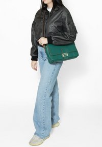 Borsa a spalla verde in pelle con forma rettangolare, accessori in oro e cuciture marroni, indossata con una giacca bomber nera e jeans blu.