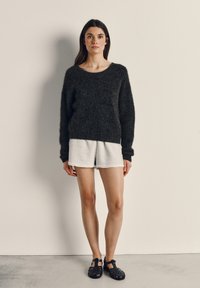 Jauna sieviete stāv, valkājot vaļīgu melnu džemperi, gaiši pelēkas šortus un melnas slēgtas sandales pret vienkāršu gaišu sienu.