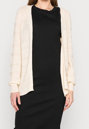 Helles beiges Cardigan mit offenem Front, gestrickter Textur und lockeren Ärmeln, getragen über einem schwarzen, gerippten Kleid.