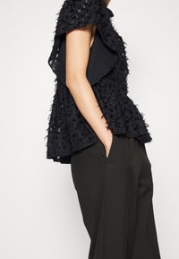 Personne portant un haut noir à manches courtes texturé avec des détails à rabat et un pantalon noir ample, la main glissée dans la poche, vue de profil.