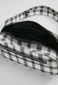 Sac à carreaux noir et blanc en matériau synthétique, doté d'une fermeture éclair et d'un intérieur doublé avec une étiquette de marque à l'intérieur.