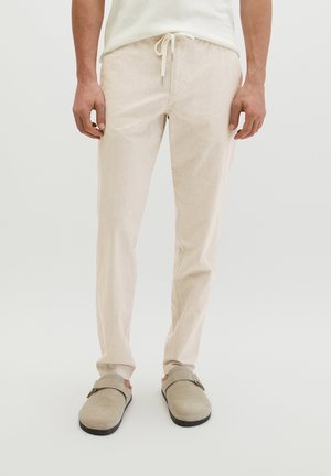 Mężczyzna w beżowych spodniach z sznurkiem i beżowych wsuwanych butach stoi na jednolitym tle.