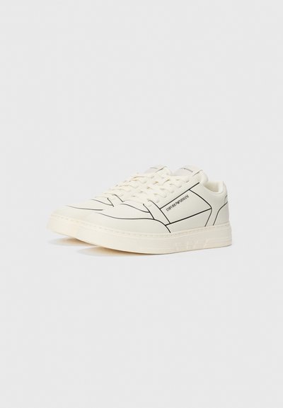 Baskets blanches avec des accents de lignes noires, en matériau lisse semblable au cuir, à bout rond, et avec une marque textuelle sur le côté, dotées d'une semelle texturée.