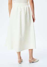 Jupe midi texturée blanche avec une taille élastique, ornée d'un motif floral. Associée à des chaussures à talons ouverts blanches.