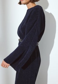 Robe plissée bleu marine avec de longues manches effilées et un col rond. Présente une ceinture métallique à la taille et un tissu texturé.