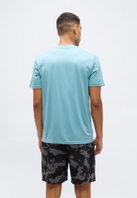 Άνδρας με κον τά μαλλιά, φοράει απλό ανοιχτό γαλάζιο κοντομάνικο πουκάμισο και σορτς καμουφλάζ σε μαύρο-γκρι, στέκεται με την πλάτη στον θεατή, μπροστά σε λευκό φόντο.