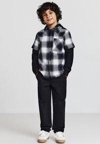 Camicia a maniche corte a quadri nera, bianca e grigia con taschino sul petto; abbinata a pantaloni neri e sneakers bianche con dettagli beige.