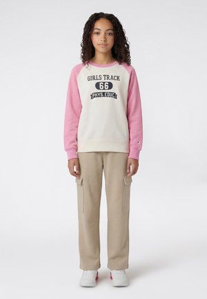 Joven de pie, con sudadera blanca y rosa con el texto "Girls Track 66 Phys. Educ.", pantalones cargo beige y zapatillas blancas.
