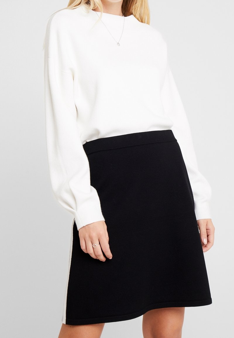 Pullover bianco a maglia con scollo rotondo e maniche lunghe abbinato a una gonna nera a trapezio con un design semplice e sottili spacchi laterali.
