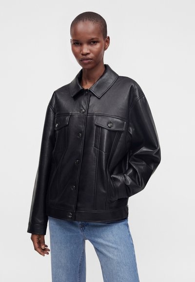 Personne aux cheveux courts portant une veste en cuir noire boutonnée et un jean bleu clair, debout avec une main dans la poche devant un fond uni.