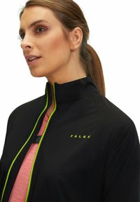 Zwarte sportjack met een hoge kraag en limoen groene ritsdetails, met het logo "FALKE" in bijpassend groen op de voorkant.