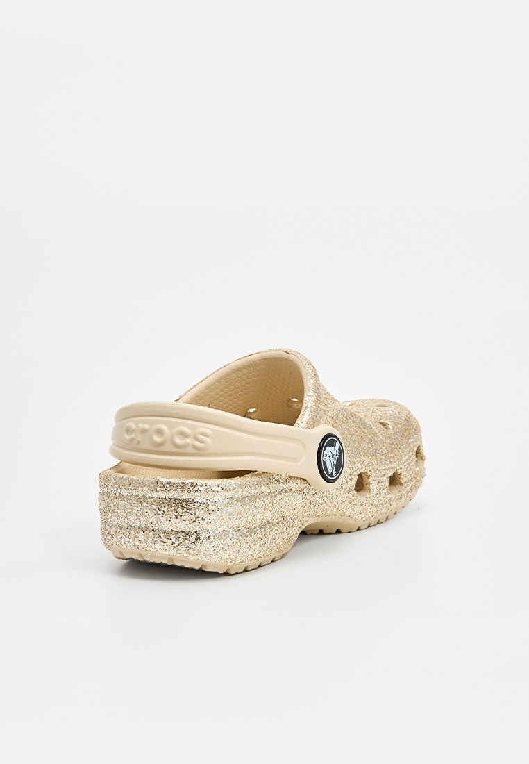 Zoccoli Crocs beige glitterati con una finitura testurizzata, cinturino posteriore, fori di ventilazione e un dettaglio logo circolare di lato.