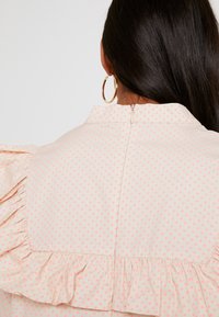 Blouse crème à pois roses, détail volanté aux épaules et fermeture éclair dans le dos. Tissu lisse avec une coupe structurée.