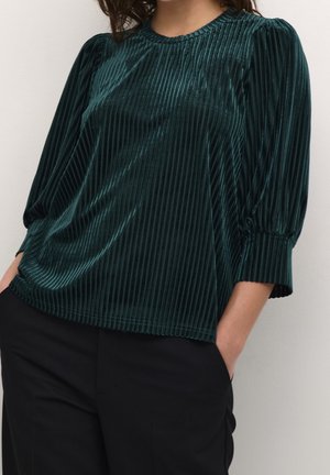 Femme portant un chemisier côtelé en velours vert foncé avec des manches bouffantes 3/4 et un pantalon noir, debout les mains dans les poches.