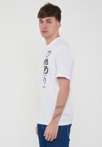 T-shirt bianco in cotone con maniche corte, presenta testo e grafica neri sul davanti, include un elemento di design blu e dettagli del logo.