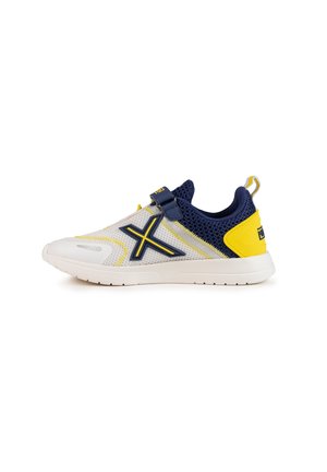 Zapatilla deportiva ligera con parte superior de malla blanca, detalles en azul marino, tacón amarillo y una correa cruzada. Presenta una superficie texturizada y suela de goma.