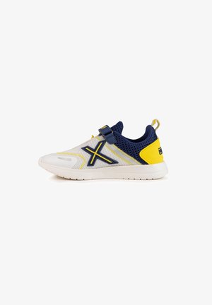 Zapatilla deportiva ligera con parte superior de malla blanca, detalles en azul marino, tacón amarillo y una correa cruzada. Presenta una superficie texturizada y suela de goma.
