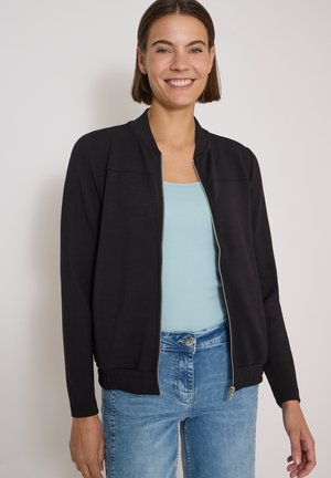 Jeune femme souriante, portant une veste zippée noire par-dessus un haut bleu clair et un jean bleu, debout devant un fond clair uni.