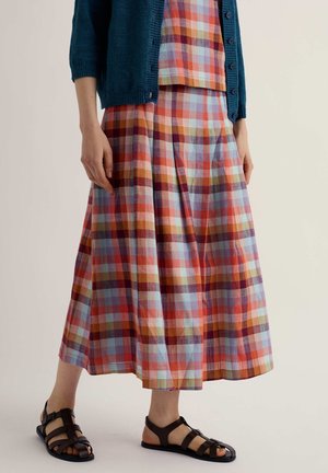 Žena oblečená v viacfarebnej kárované sukni a topi s tyrkysovým kardigánom a čiernymi sandálmi na remienky, stojaca pred jednofarebným pozadím.