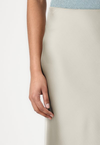 Jupe beige clair en matériau lisse, avec un design ajusté et un éclat subtil. Une main effleure légèrement le bord du tissu.