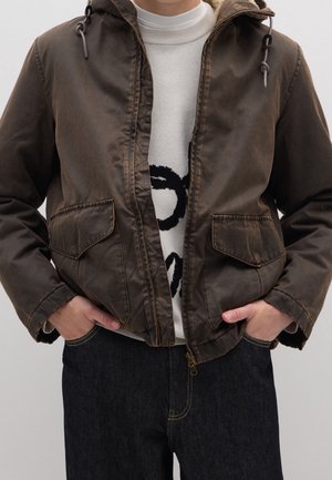 Person iført en brun hættejakke over en hvid sweater med sort tekst, hænderne i jakkelommerne, kombineret med mørke denimjeans.