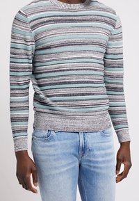 Gestreifter Pullover in Grau, Hellblau und Schwarz. Runder Halsausschnitt, gerippte Bündchen und Saum. Über hellblauen Jeans getragen. Glatte Textur.