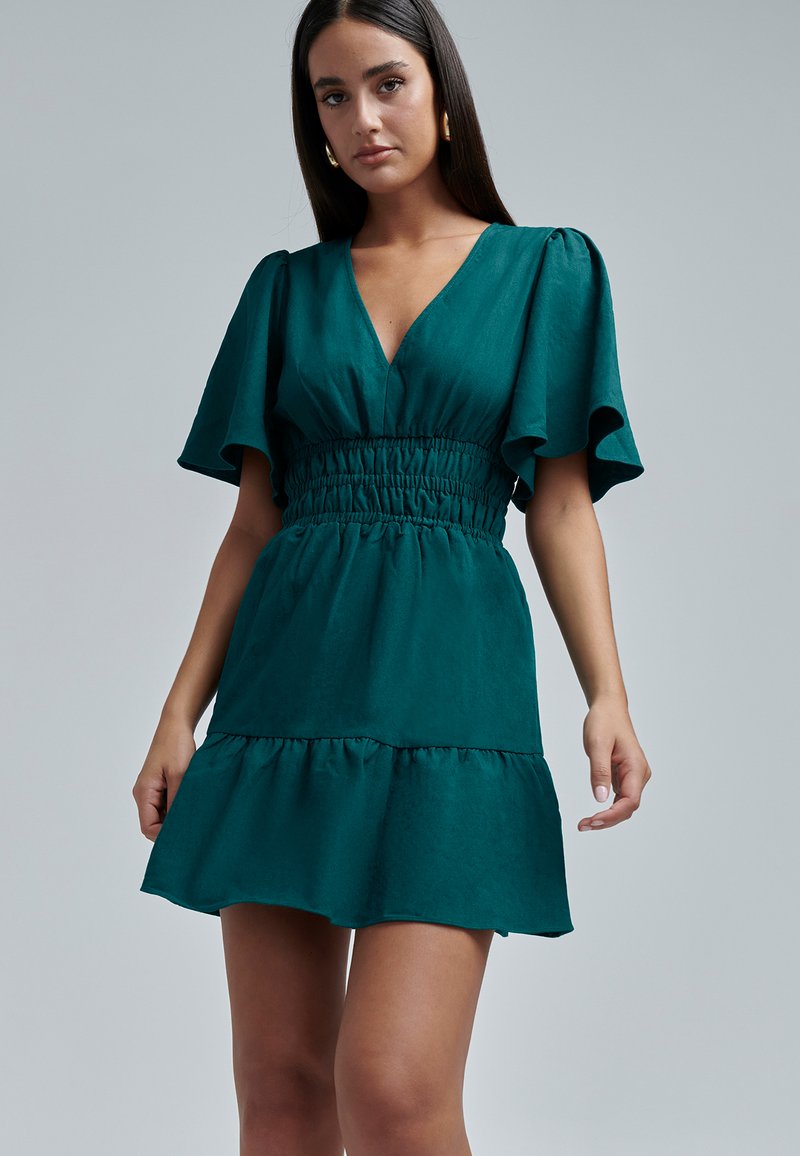 Teal lyhyt mekko, jossa on ryhmittelyvyötärö, v-mallinen kaulus ja liehuvat hihat. Siinä on kerroksellinen hame ja pehmeä, teksturoitu kangas.