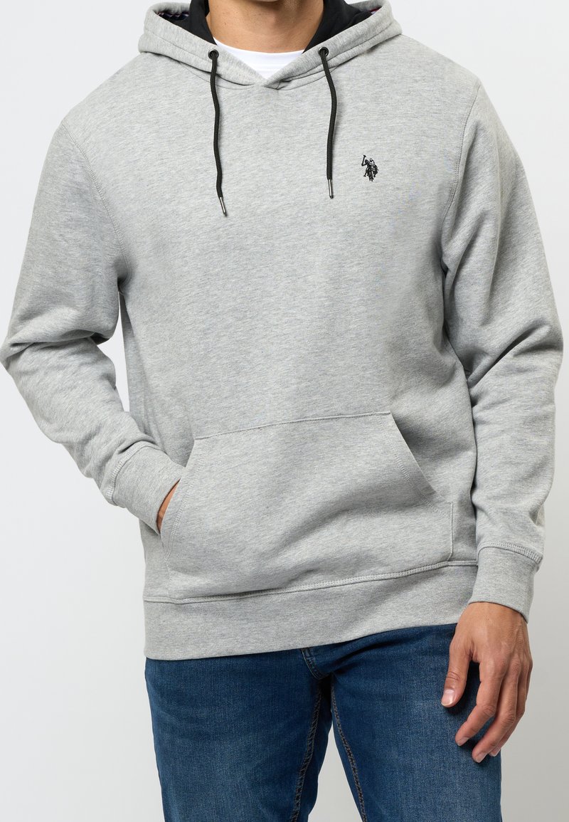 Homme portant un sweat à capuche gris clair avec cordons noirs et poche kangourou, associé à un jean bleu, main dans la poche, torse visible.