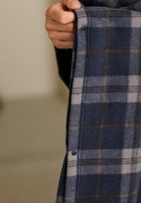 Marineblaues karierter Flanellhemd mit hellgrauen und braunen Linien; verfügt über einen schwarzen Druckknopfdetail entlang des Saums; weicher, strukturierter Stoff.