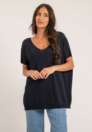 Femme aux longs cheveux noirs portant un haut noir à manches larges et col en V ainsi qu'un jean bleu clair, debout devant un fond uni.